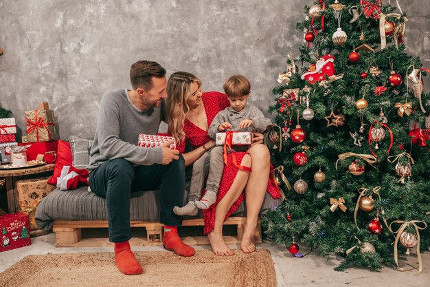 La famiglia con un bambino si diverte con una confezione regalo vicino all'albero di Natale sul divano, colori rosso verde, decorazioni per la casa, momenti felici. Coppia con figlio