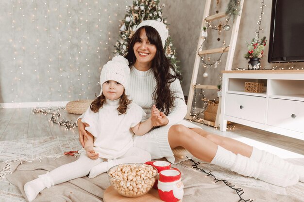 La famiglia felice, la madre e la bambina con cappelli a maglia bianchi siedono vicino a un albero di Natale con un regalo di Natale