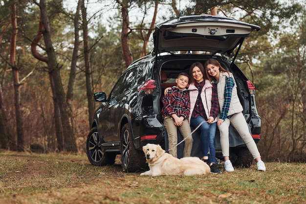 La famiglia felice si diverte con il proprio cane vicino a un'auto moderna all'aperto nella foresta