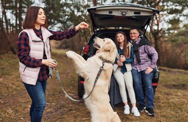 La famiglia felice si diverte con il proprio cane vicino a un'auto moderna all'aperto nella foresta