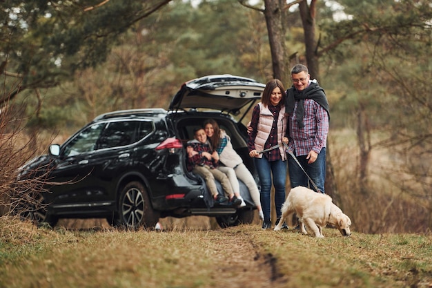 La famiglia felice si diverte con il proprio cane vicino a un'auto moderna all'aperto nella foresta
