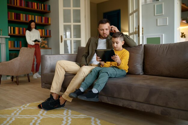 La famiglia felice si diverte insieme nel fine settimana a casa. Padre e figlio che si abbracciano durante l'utilizzo del tablet mobile mentre la madre legge il libro