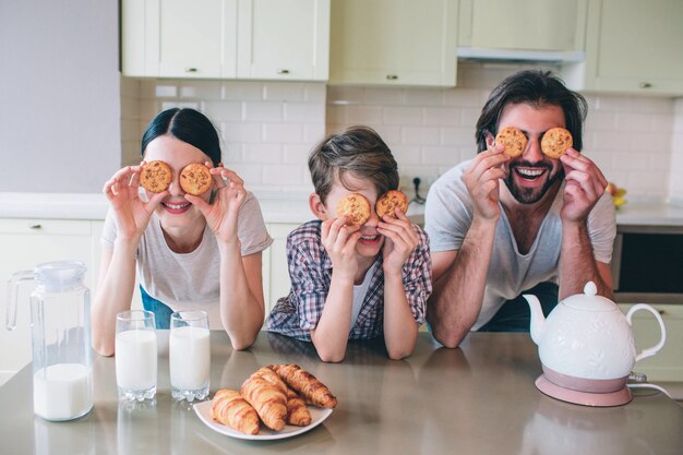 La famiglia felice sta giocando con i panini. Li pitano sugli occhi e sorridono. La famiglia è appoggiata al tavolo. Ci sono bollitore, latte e cornetti.