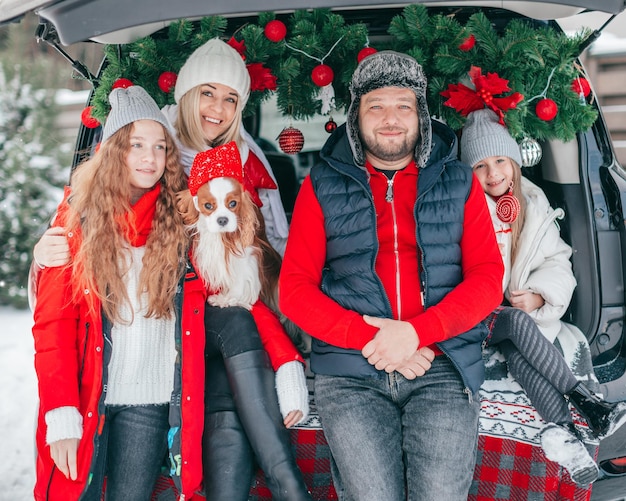 La famiglia si diverte nel bagagliaio dell'auto. Padre madre due figlie felici di trascorrere del tempo insieme. Vacanze invernali Natale. Decorazione rossa. Vigilia di Capodanno