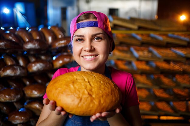 La fase di cottura in una panetteria. Ritratto di una ragazza fornaio con pane in mano sullo sfondo di scaffalature in una panetteria. Mani di un fornaio con pane. Produzione di pane industriale