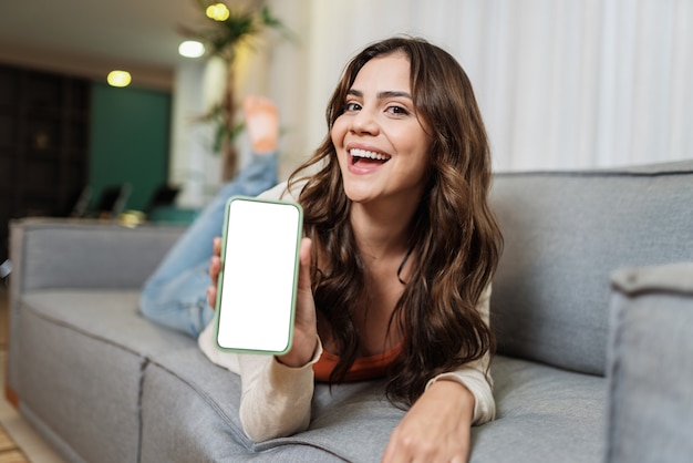 La felice signora latina si rilassa a casa da sola nella posa della stanza e condivide buone notizie sui social media tramite il cellulare. Donna sorridente che mostra l'app del telefono.