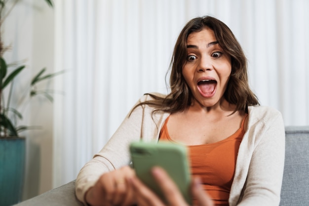 La felice signora latina si rilassa a casa da sola, si siede sulla posa della stanza e condivide buone notizie sui social media tramite il cellulare. La donna sorridente si gode il cibo per gli ordini del fine settimana online nell'app del telefono.