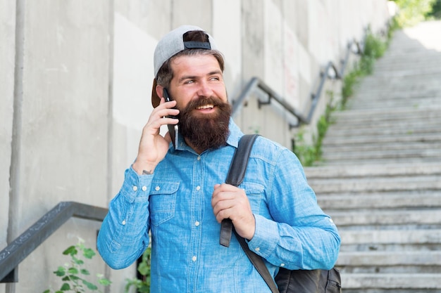 La felicità è viaggiare Uomo barbuto parlare al telefono felice uomo brutale a piedi strada Barbiere maschio cura pantaloni a vita bassa brutali con zaino escursionismo avventura concetto stile urbano Viaggiatore hipster maturo