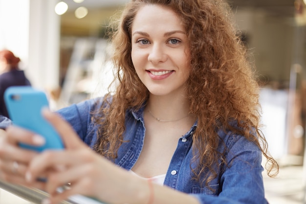 La femmina abbastanza giovane tiene lo Smart Phone, utilizza la connessione Internet gratuita nel centro commerciale, i messaggi con gli amici, sta al coperto