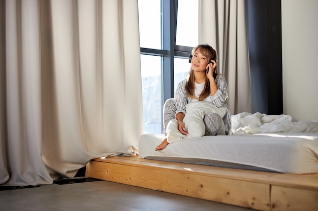 La femmina asiatica gode della musica in cuffie che si siede sul letto, donna felice in pigiama si siede da solo in camera da letto