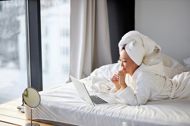 La femmina asiatica guarda lo schermo del laptop parlando, sorridendo, conversando online con qualcuno, a casa sul letto