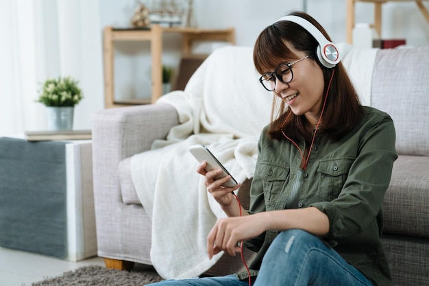 la femmina asiatica seduta e sorridente sta sfogliando le informazioni sul suo telefono nel soggiorno. la signora che indossa gli auricolari con uno stato d'animo rilassato ascolta canzoni e fa acquisti online nel suo tempo libero.