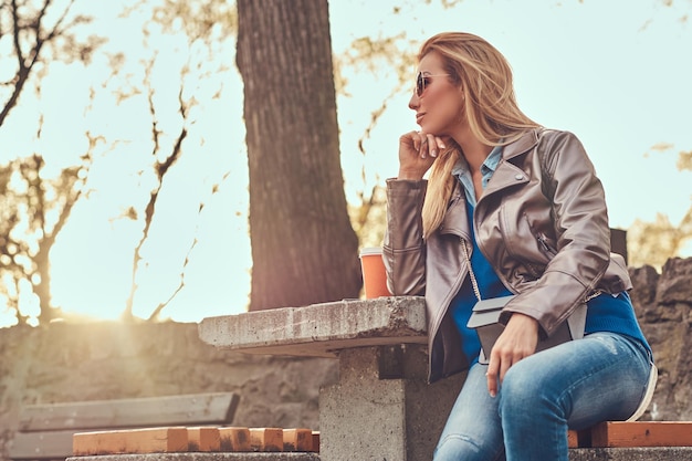 La femmina bionda alla moda si rilassa all'aperto, sedendosi sulla panchina nel parco cittadino contro una luce solare intensa.