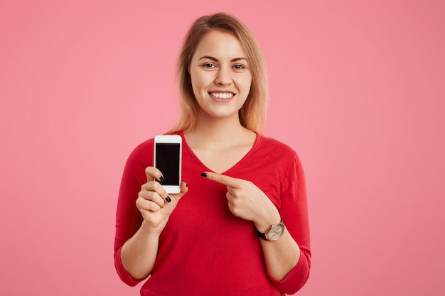 La femmina bionda allegra con l'ampio sorriso indica allo schermo in bianco dello Smart Phone per il vostro contenuto pubblicitario, vestita in maglione rosso, isolata sulla parete rosa. Concetto di tecnologia e persone