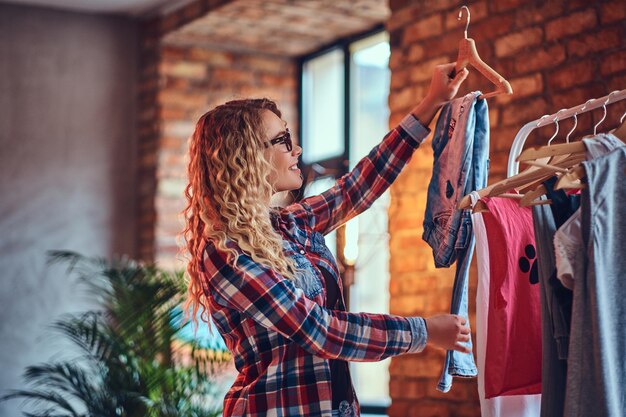 La femmina bionda positiva con gli occhiali neri sceglie i vestiti alla moda sull'attaccapanni.