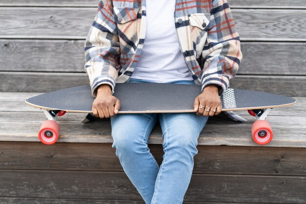 La femmina etnica africana in abiti casual tiene il longboard seduto su una panca di legno con lo skateboard in mano