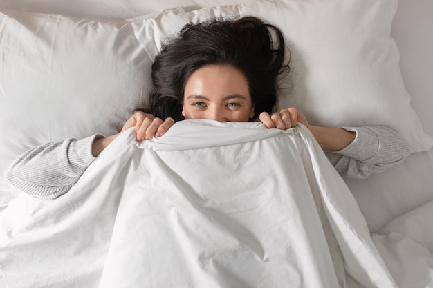 La femmina europea abbastanza giovane e felice dorme sul letto bianco copre il viso con la coperta si sveglia gode del buongiorno