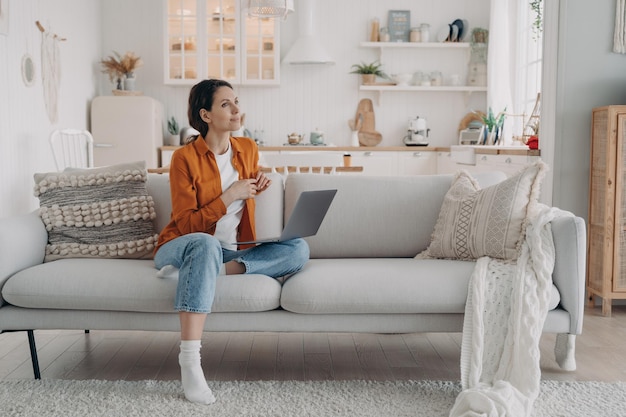 La femmina sognante pensierosa riposa con il laptop su un comodo divano moderno nell'accogliente soggiorno di casa