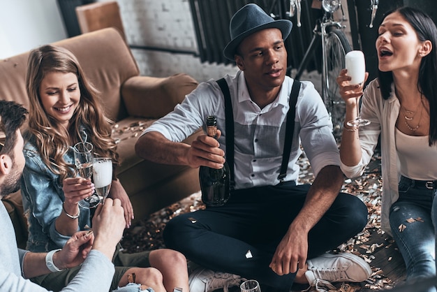 La festa è impossibile senza champagne! Vista dall'alto dei giovani moderni che bevono champagne seduti sul pavimento con i coriandoli intorno