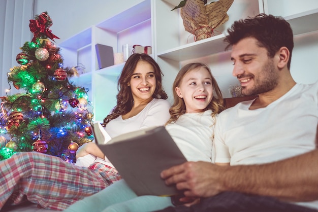 La figlia e i genitori felici si sdraiano su un letto e leggono un libro. sera notte