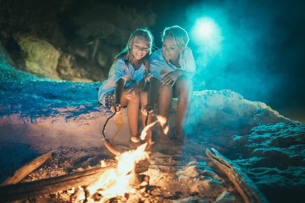 La figlia sorridente felice e sua madre sono sedute sulla spiaggia sabbiosa vicino al fuoco la sera. Stanno friggendo le salsicce sul bastone.