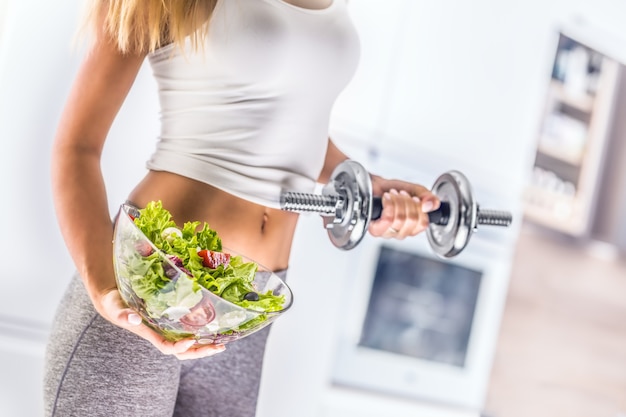 La figura sportiva di una donna attraente che tiene in mano una ciotola di insalata di verdure fresche e manubri.