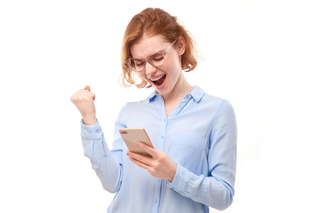 La fortunata ragazza rossa sorridente che tiene in mano uno smartphone in camicia da lavoro con occhiali isolati in studio ha ricevuto un messaggio gioioso, celebrando la vittoria