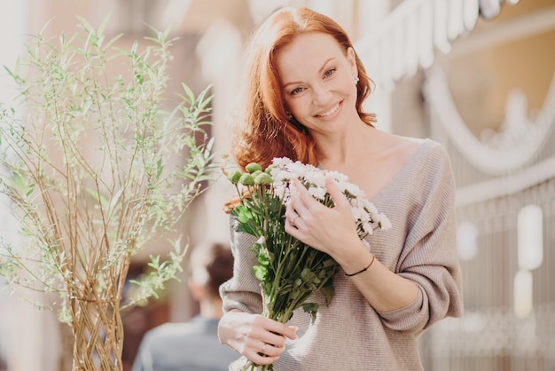 La foto all'aperto della bella donna foxy inclina la testa felice di ricevere fiori dal fidanzato vestito con abiti casual ha appuntamento con il fidanzato Attraente donna dai capelli rossi ama la primavera Street style