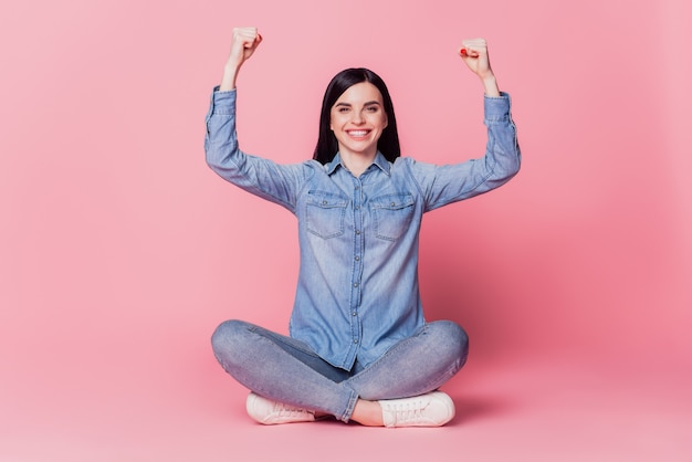 La foto completa del corpo di una ragazza allegra celebra il suo successo con i pugni le mani si siedono sul pavimento isolato su uno sfondo rosa dello studio