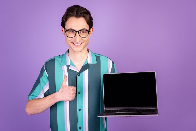 La foto del giovane mostra il pollice in alto pubblicizza la scelta approva il computer portatile della tenuta isolato su sfondo di colore viola