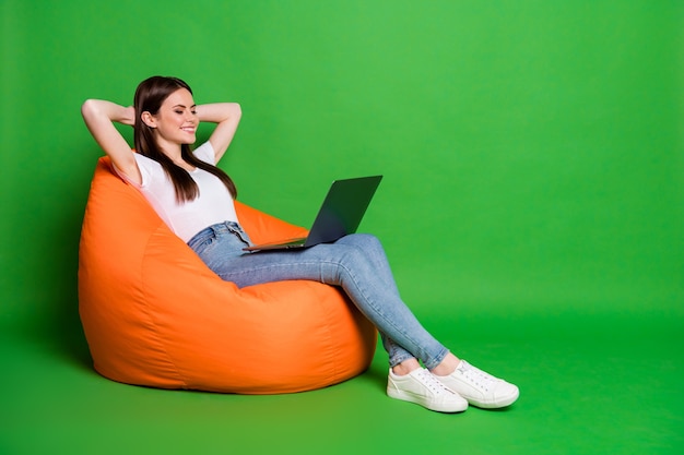 La foto del profilo di una giovane donna piuttosto allegra tiene le mani del laptop dietro la testa vestito t-shirt bianca jeans scarpe da ginnastica isolate su sfondo verde