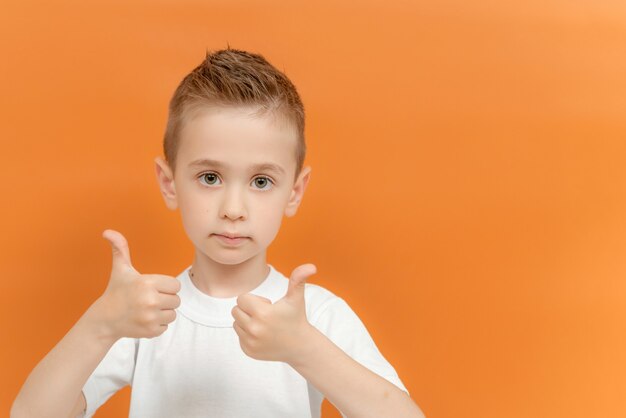 La foto del ragazzo prescolare positivo mostra il pollice in su segno di usura con due mani isolate su colore arancione