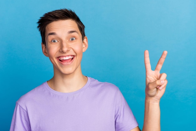 La foto del sorriso positivo felice del giovane mostra il vsign fresco di pace isolato sopra priorità bassa di colore blu