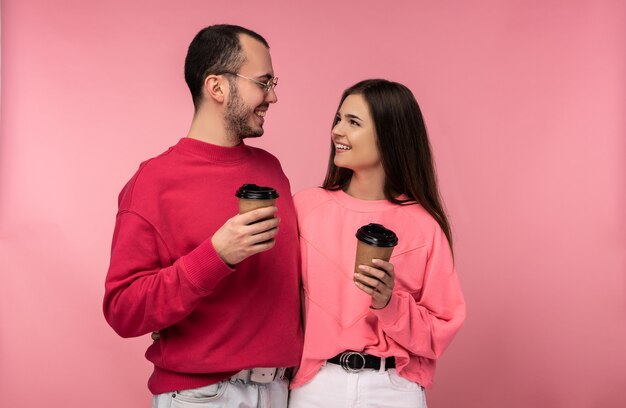 La foto dell'uomo attraente con la barba in vestiti rossi e la donna in rosa tiene caffè e gawp l'uno sull'altro. La coppia sembra felice, isolata su sfondo rosa.