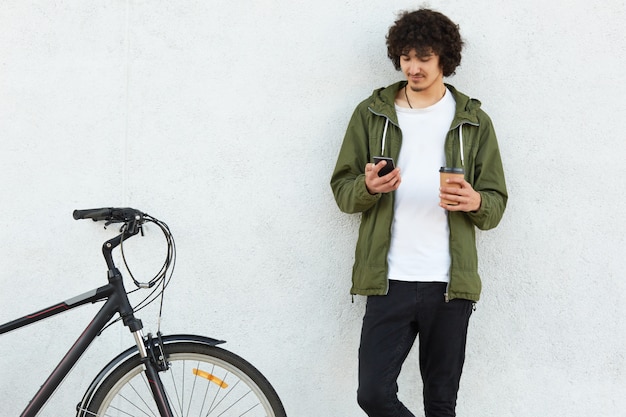 La foto dell'uomo attraente indossa una giacca a vento verde alla moda e jeans neri, ha la barba piccola, tiene in mano il cellulare