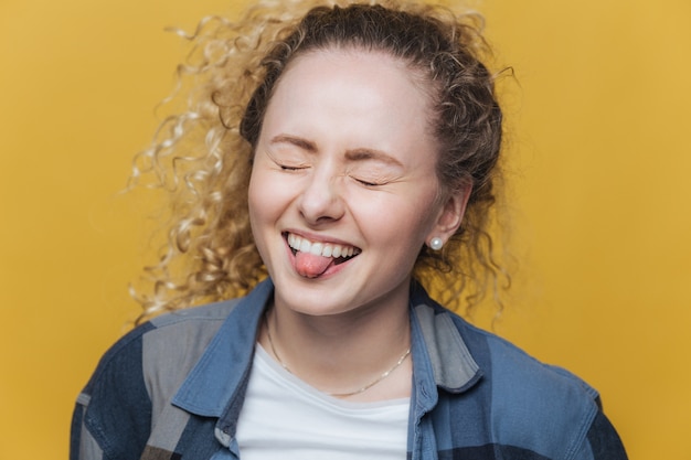 La foto della femmina divertente e allegra si diverte da sola, ride con gioia e mostra la lingua, tiene gli occhi chiusi