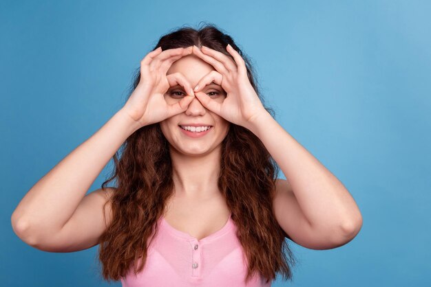 La foto della giovane donna eccitata si diverte a mostrare le dita binocolo occhiali da vista isolate su sfondo blu.