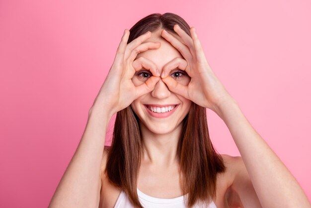 La foto della ragazza adorabile della gioventù si diverte mostra gli occhiali del binocolo delle dita isolati su uno sfondo di colore rosa