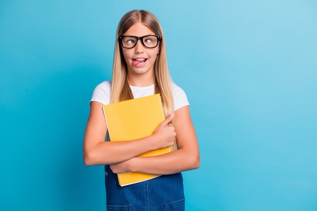 La foto della ragazza divertente dell'allievo non vuole fare i compiti mostra la lingua indossare gli occhiali tenere il libro isolato su uno sfondo di colore blu pastello