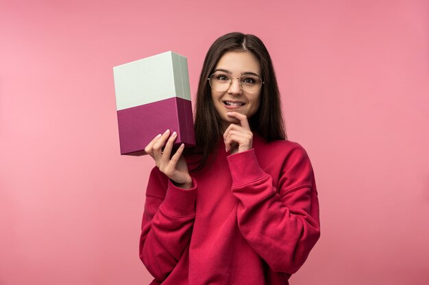 La foto della signora attraente con gli occhiali tiene la scatola con il presente, attende e non vede l'ora di aprire. Indossa pantaloni bianchi casual maglione rosa isolato sfondo di colore rosa.