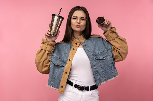 La foto della signora attraente tiene i muffin del caffè del tè felici mostra l'amore, indossa la camicia dei jeans casuali la maglietta bianca isolata il fondo di colore rosa.