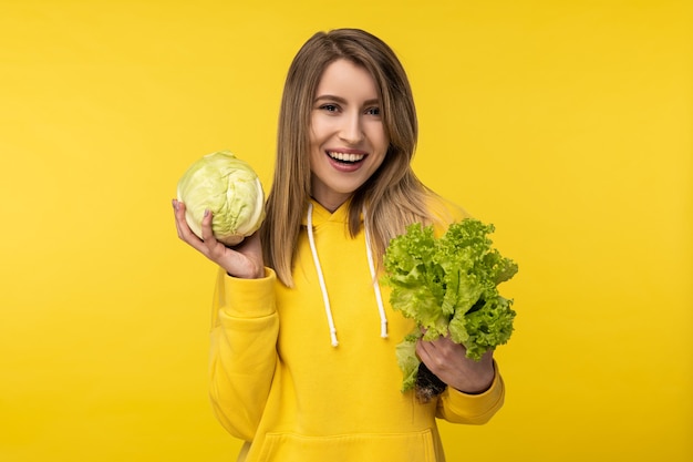 La foto della signora attraente tiene il cavolo e l'insalata, felice del cibo sano. Indossa una felpa con cappuccio gialla casual, sfondo di colore giallo isolato.