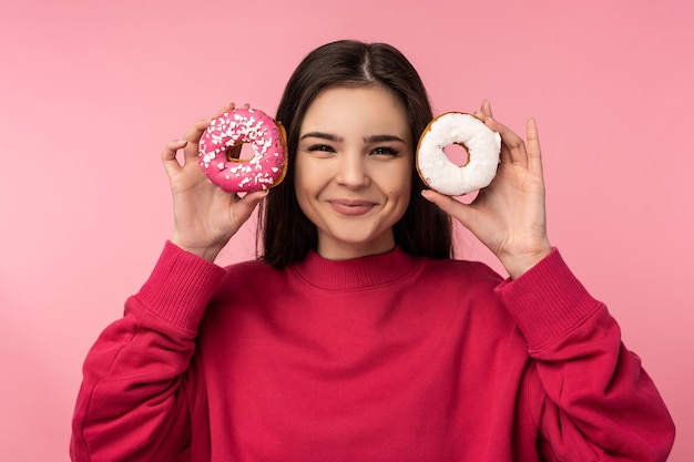 La foto della signora attraente tiene le caramelle della ciambella l'usura del cibo dolce sorriso felice maglione rosa casuale isolato sfondo di colore rosa.