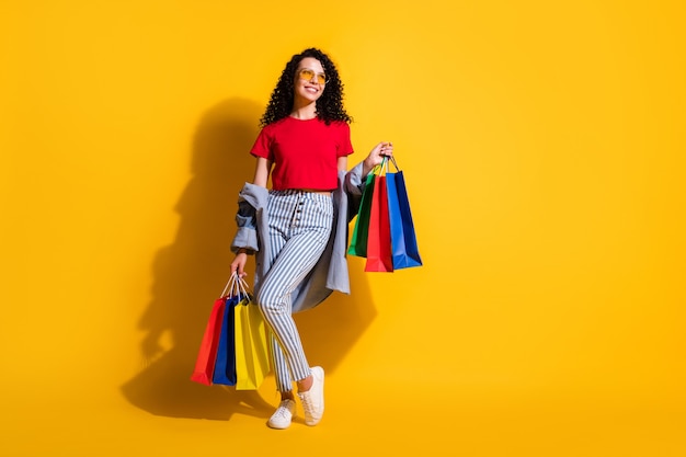 La foto della signora cerca lo spazio vuoto tiene i pacchetti del negozio indossa una t-shirt rossa jeans a righe giacca occhiali da sole sneakers isolato colore giallo sfondo