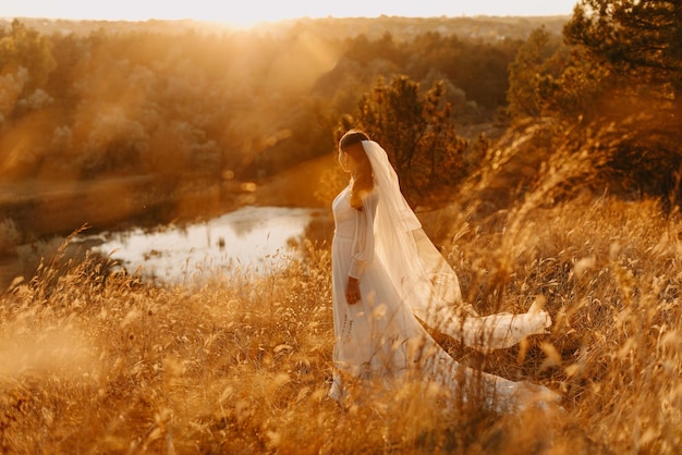 La foto della sposa sognante in un vestito lussureggiante gira vicino al lago al tramonto