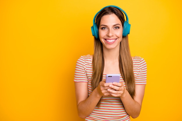 La foto di bella signora che tiene il telefono ascolta musica in auricolari wireless fantastici scegliendo la prossima canzone indossare maglietta a righe casual isolata parete di colore giallo