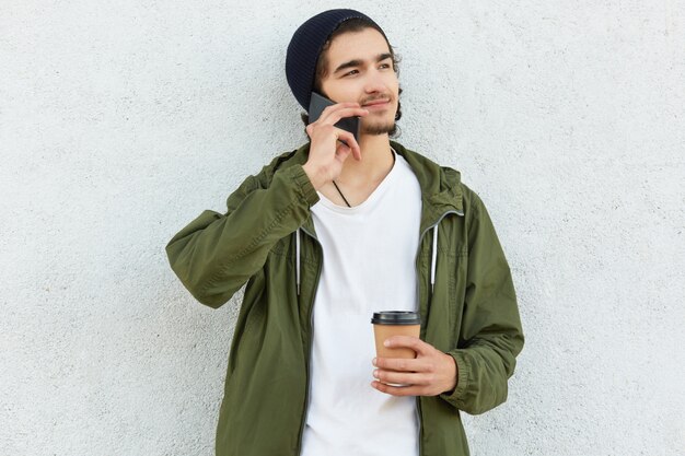 La foto di hipster felice sembra pensierosa a parte, beve caffè aromatico, parla con qualcuno tramite smartphone