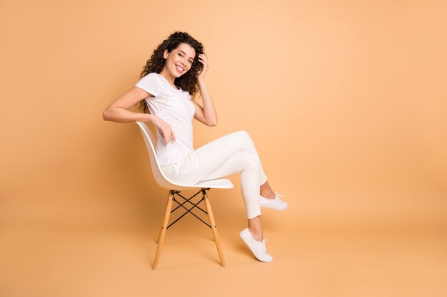 La foto di profilo a grandezza naturale della signora di affari incredibile che si siede sulla sedia comoda che sorride le forme sottili positive indossano i vestiti casuali isolati il fondo di colore pastello beige