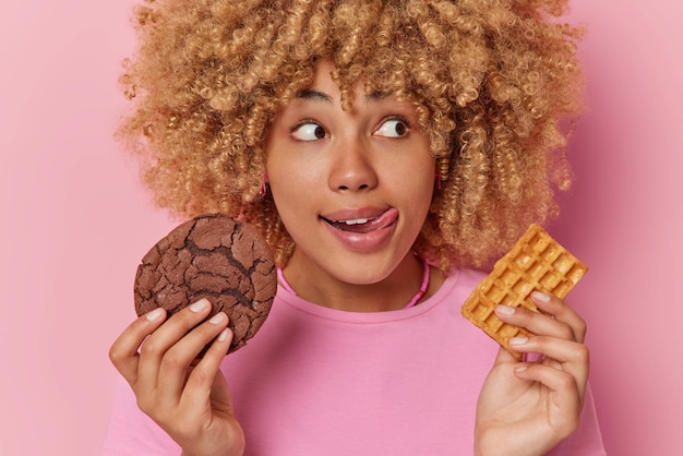 La foto di un adorabile modello femminile con i capelli ricci si lecca le labbra tiene un biscotto appetitoso e la cialda gode di uno spuntino ha un debole per i dolci indossa una maglietta casual isolata su sfondo rosa Concetto di cibo malsano