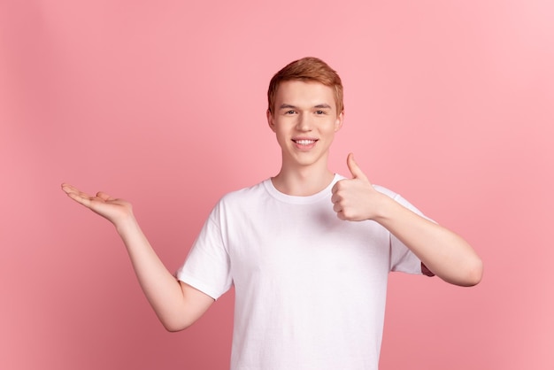 La foto di un giovane allegro promotore tiene la proposta del braccio mostra il pollice in su suggerisce la qualità isolata su uno sfondo di colore rosa